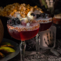 Two dark red Black Widow (Kit) cocktails in coupe glasses rimmed with brown sugar, garnished with blackberries and lychee, sit by smoky vapor, a skeleton hand candle, and popcorn—a mysterious cinnamon plum cocktail kit experience.