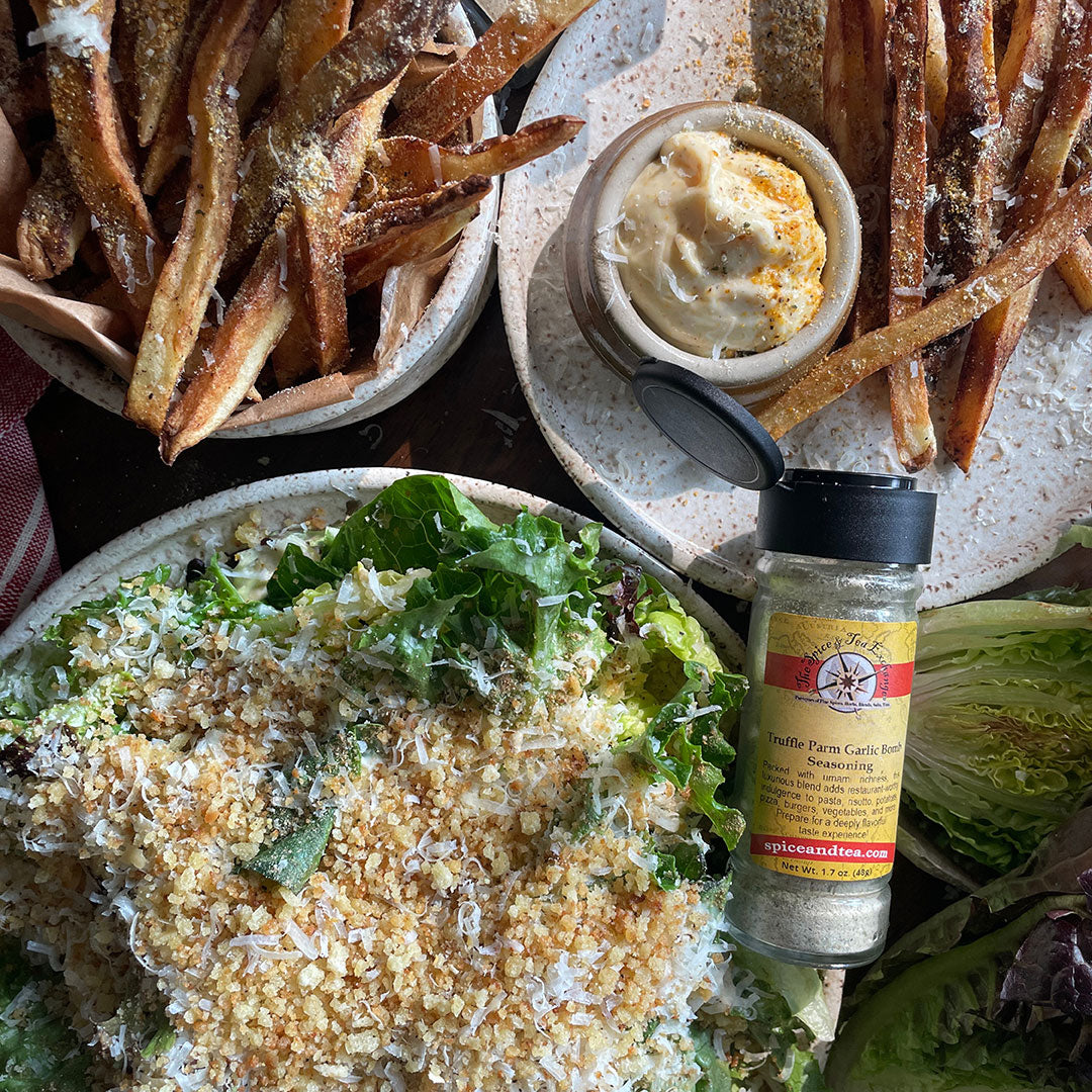 Plates of French fries with dipping sauce, a leafy green salad topped with breadcrumbs and grated cheese, and a jar of seasoning blend on a rustic table.