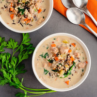 Two white bowls of creamy chicken and rice soup with vegetables, arranged on a gray surface with parsley and silver spoons on an orange napkin.