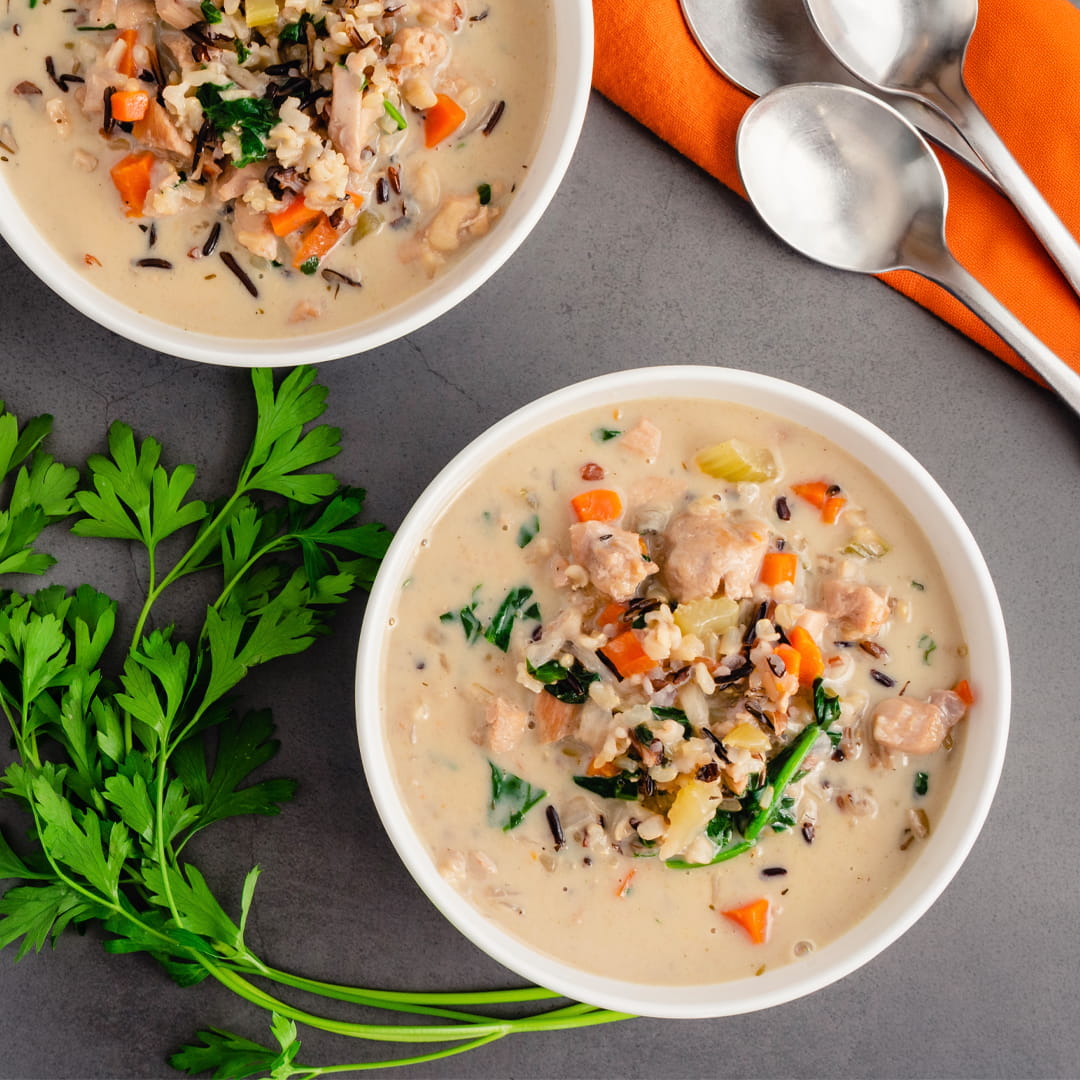Two white bowls of creamy chicken and rice soup with vegetables, arranged on a gray surface with parsley and silver spoons on an orange napkin.