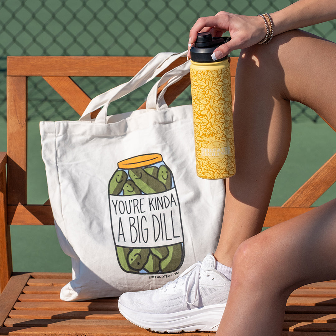 You're Kinda A Big Dill Tote Bag next to Yellow Tea Leaf Tumbler on pickleball court bench