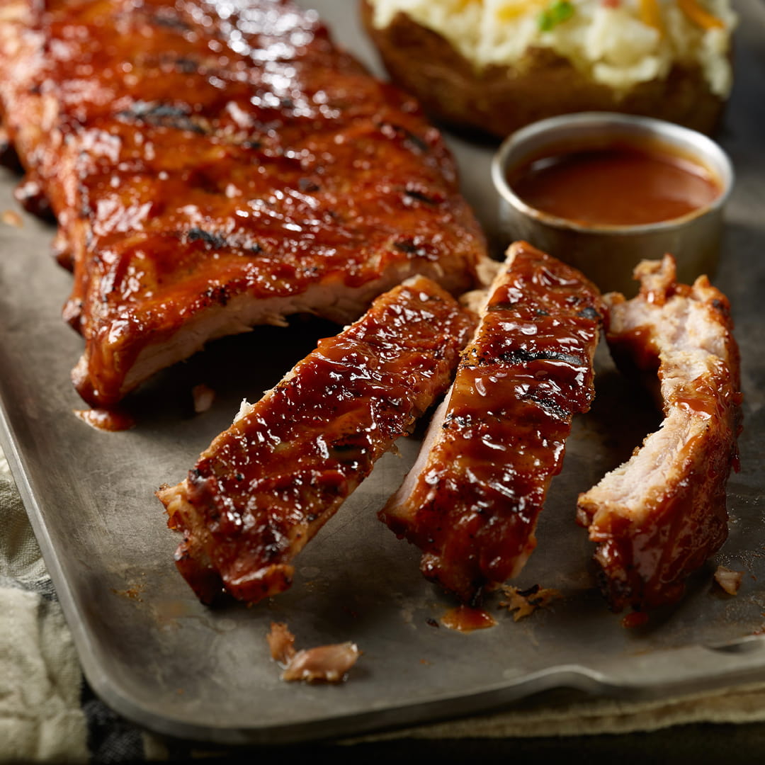 Succulent glazed pork ribs served on a metal tray with sliced pieces, a baked potato, and a small cup of red dipping sauce.
