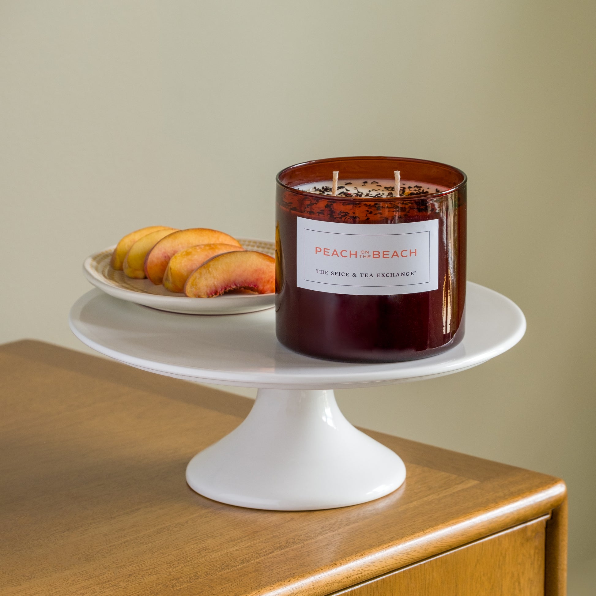 A brown glass Peach on the Beach Candle sits on a white cake stand beside a small plate of sliced peaches, all arranged on a wooden surface with a neutral background.