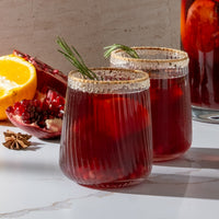 Two ribbed clear glass tumblers filled with a deep red beverage, rimmed with sugar, and garnished with a sprig of rosemary, set on a light marble surface with pomegranate, orange, and star anise nearby.